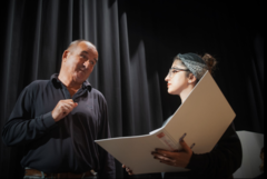Der Regisseur und Theaterdirektor Manfred Michalke steht auf einer Bühne neben einer jungen Frau, die eine offene weiße Mappe in der Hand hält. Sie haben Augenkontakt.
