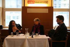 Um einen Tisch mit weißem Tischtuch sitzen links und in der Mitte eine Frau und rechts ein Mann mit Headsets. Aufschrift: ORF-Radiocafe