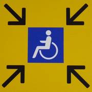Rollstuhlzeichen vor gelbem Hintergrund; in den Ecken sind jeweils Pfeile, die auf den Rollstuhl hinweisen