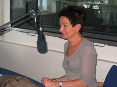eine Frau mit schwarzen Kurzen Haaren in einem grauen Kostum sitzt vor einem Mikrophon in einem Studio