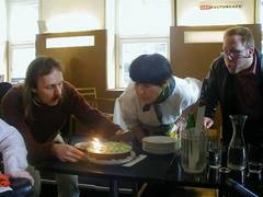 Ein Mann mit langen Haaren und Bart und eine Frau mit schwarzen Haar schauen eine Torte mit brennender Kerze an