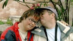 Eine Frau mit Behinderung im roten Anorak wird von einem Mann mit Behinderung mit Kappe geküsst