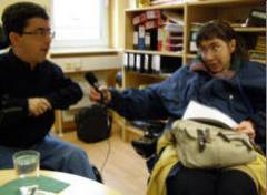 Ein Rollstuhlfahrer mit dunklem Haar und Brille wird von einer Rollstuhlfahrerin mit brünettem, längerem Haar und Brille interviewt.