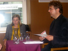 Bild 1: Eine Frau und ein Mann sitzen an einem Tisch auf einem Podium. Der Mann hält ein Papier in der Hand und redet.