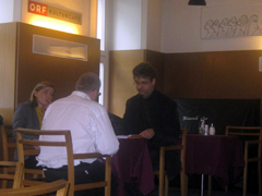 Bild 2: Drei Personen sitzen um einen Tisch auf dem Podium des KulturCafes und diskutieren.